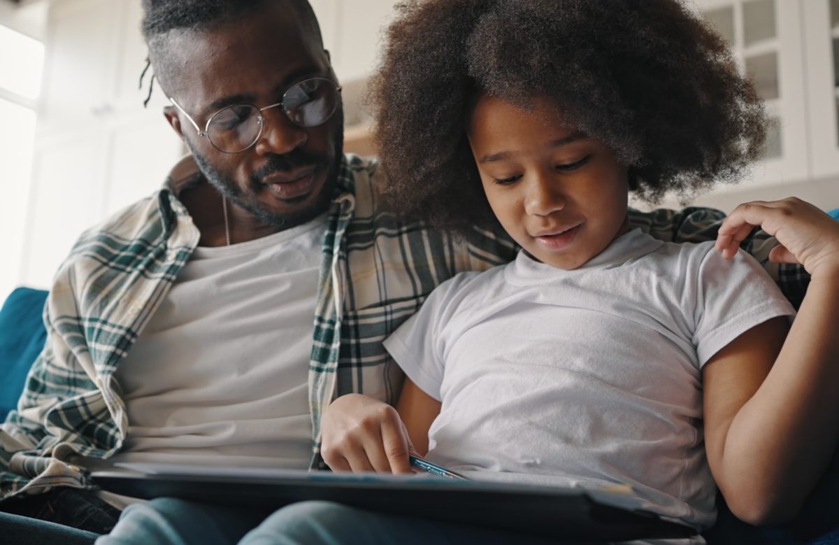 A father and his daughter sitting together on a sofa, looking at a tablet as she points to the screen