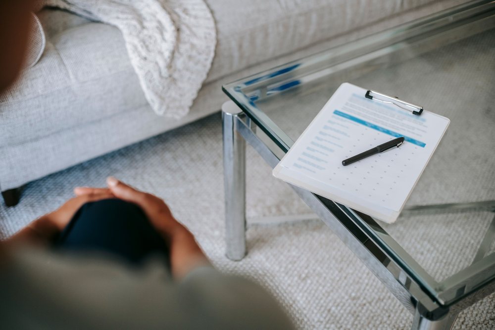 A clipboard with a questionnaire resting on a glass coffee table, with a person seated nearby on a sofa